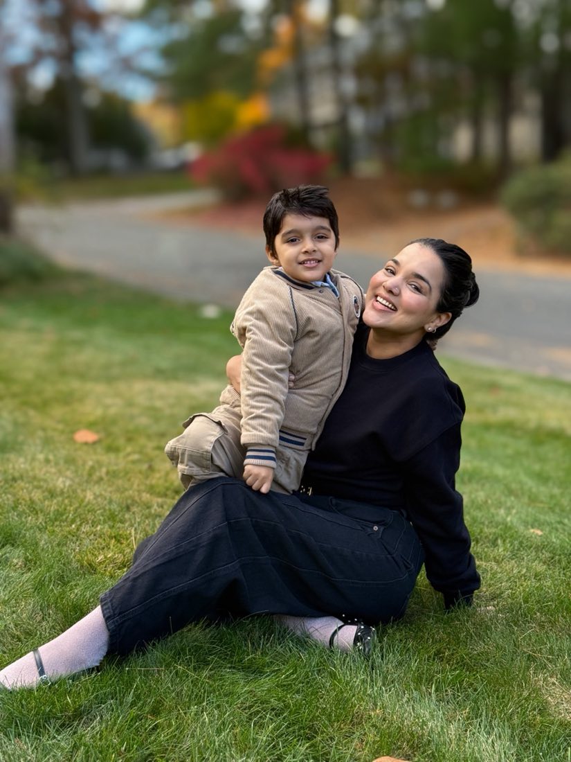 Nandita sitting on the grass with her son Yuvi on her lap, both smiling, autumn trees behind them