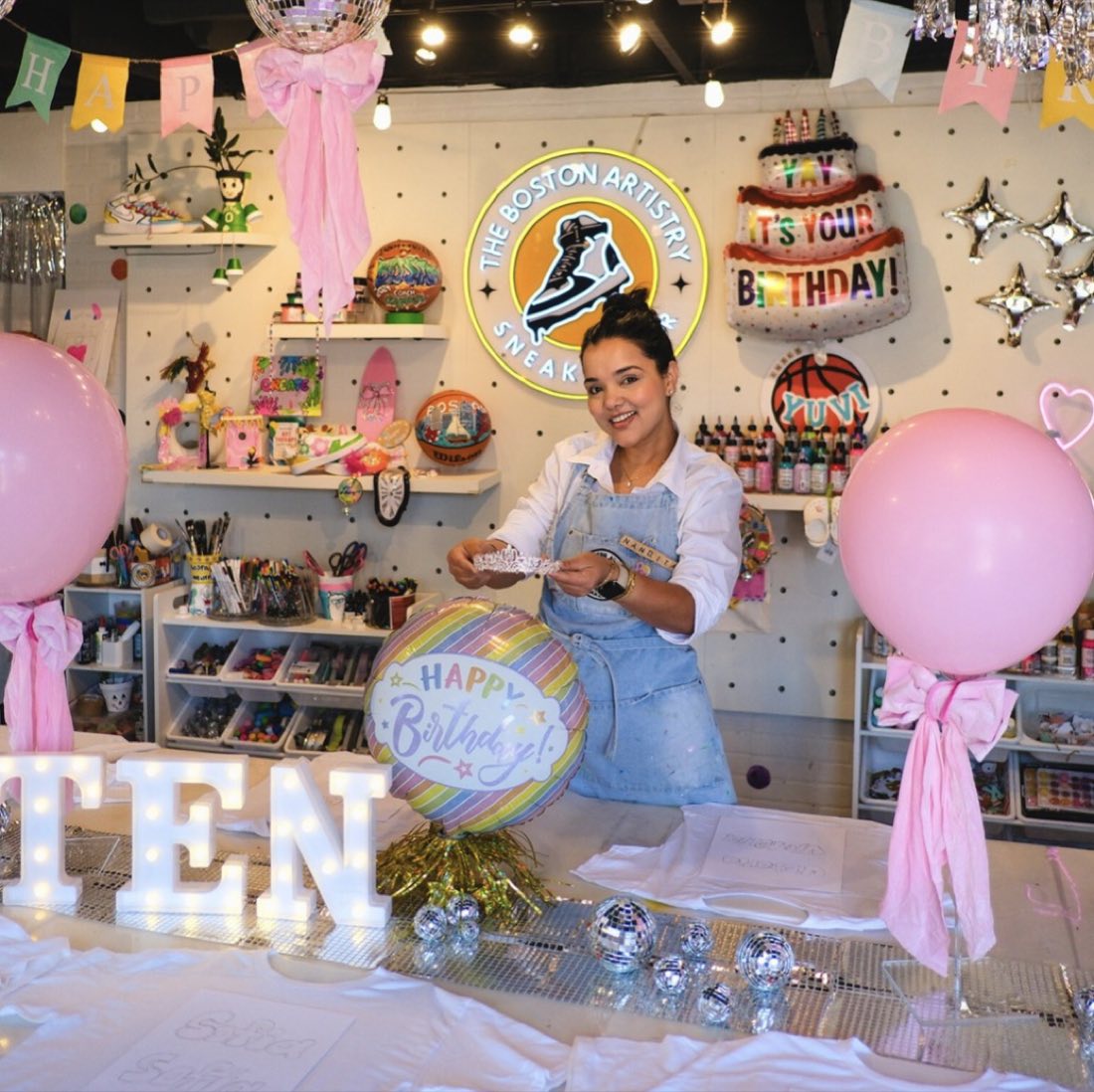 Nandita at a birthday party table in the studio, pink balloons and the Sneaker Bar sign behind her