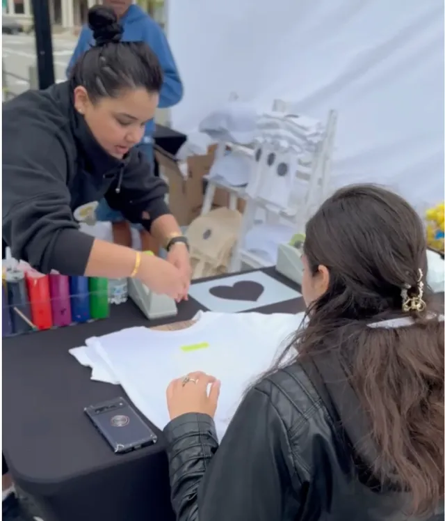 A young artist presses a heart stencil onto a shirt at an event table under a tent