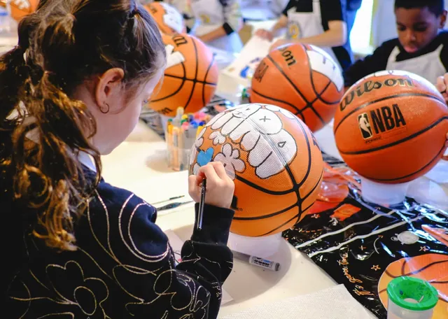 A girl paints a white design on a basketball, with more basketballs waiting on stands around her