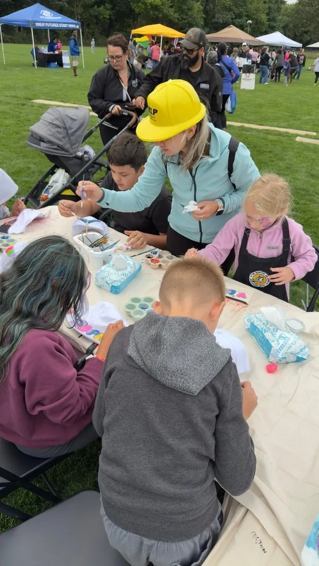 An instructor in a yellow hard hat helps kids at a craft table on the grass at an outdoor event