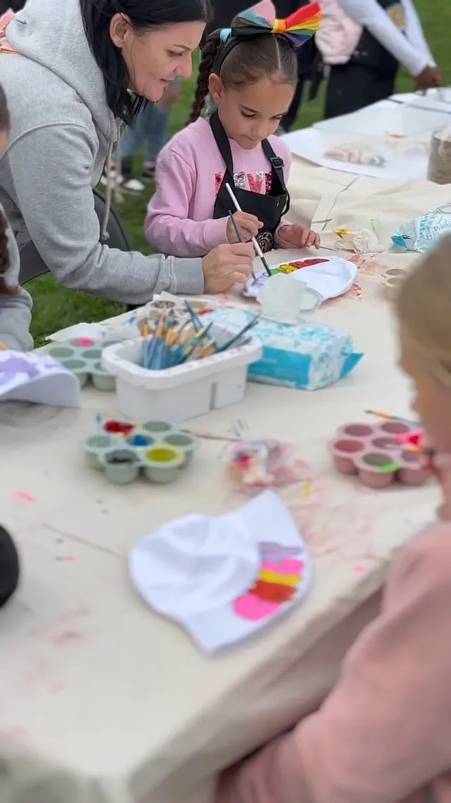 A helper guides a girl with a rainbow bow as she paints at an outdoor table on the grass