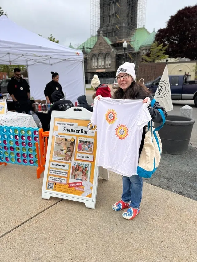 A girl in a white beanie holds up the t-shirt she painted beside The Boston Artistry Sneaker Bar sign at an outdoor pop-up