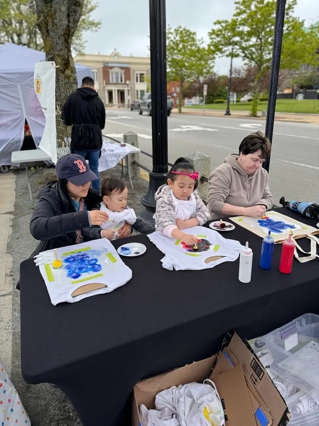 A family and a girl paint canvas tote bags at a street side pop-up table beside a white tent