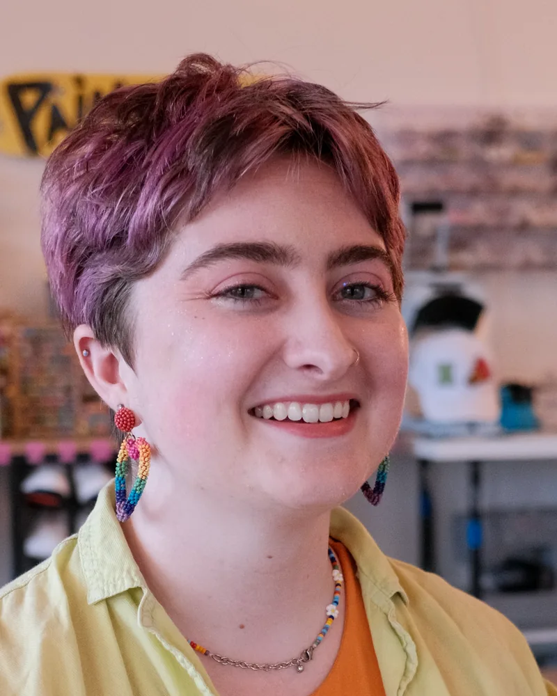 Ashley, studio artist and party host at The Boston Artistry, smiling with short hair and rainbow bead earrings in the studio