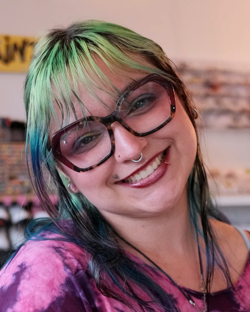 Casey, studio artist and instructor at The Boston Artistry, smiling in glasses and a tie dye T-shirt in the studio