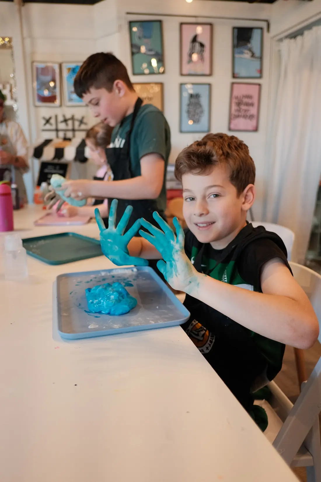 A smiling boy holds up hands covered in blue slime at the long studio table