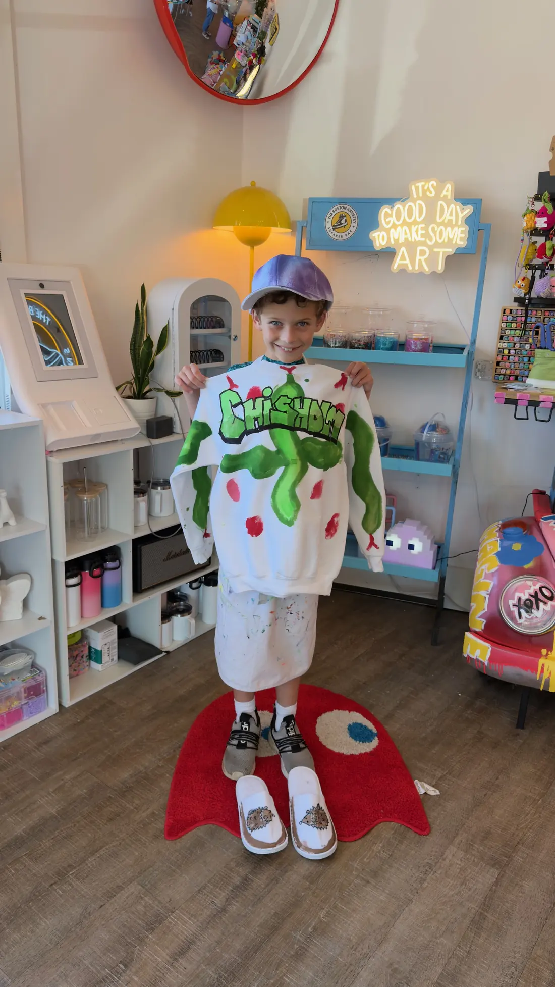 A boy in a painted purple cap holds up a white sweatshirt painted with green graffiti letters, painted shoes on the floor