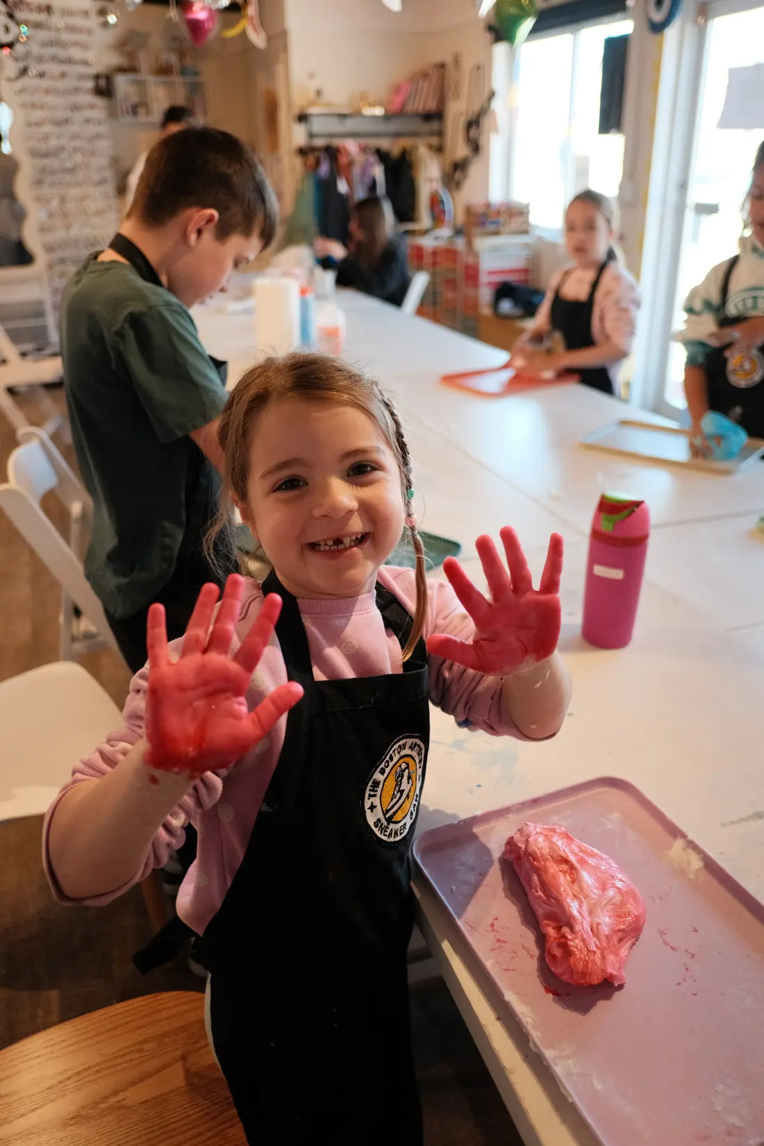 A laughing girl in a studio apron shows both hands covered in pink slime at the long studio table