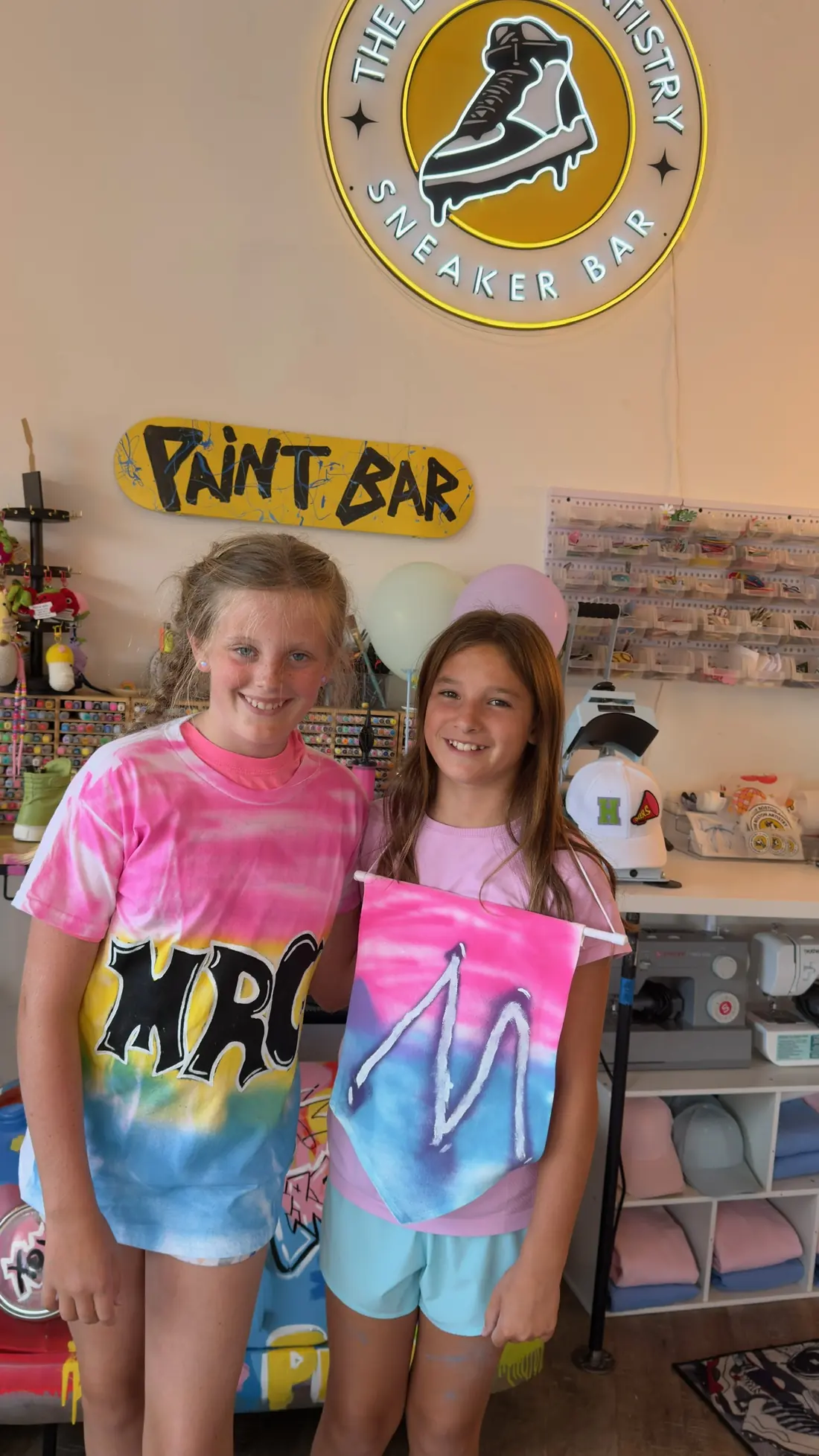Two smiling friends under the studio neon sign, one in a lettered tie dye T-shirt, one holding a tie dye pennant