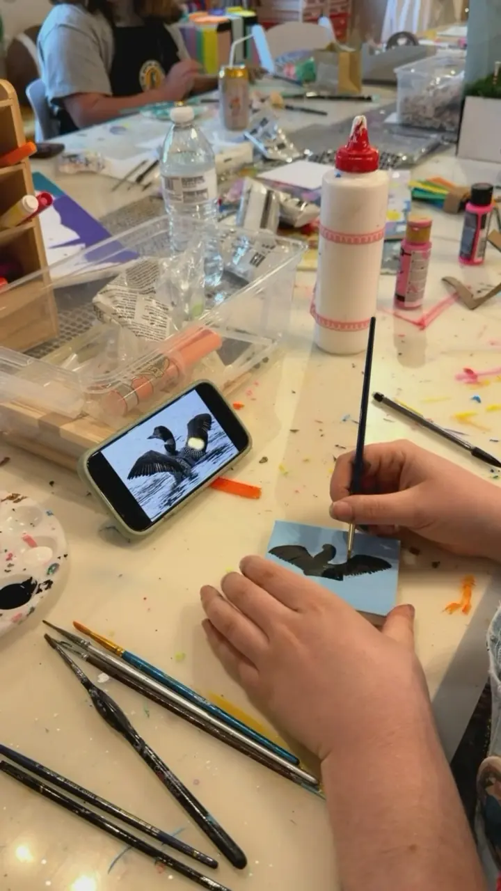 Hands paint a black bird with spread wings on a tiny blue canvas, copying a photo on a phone