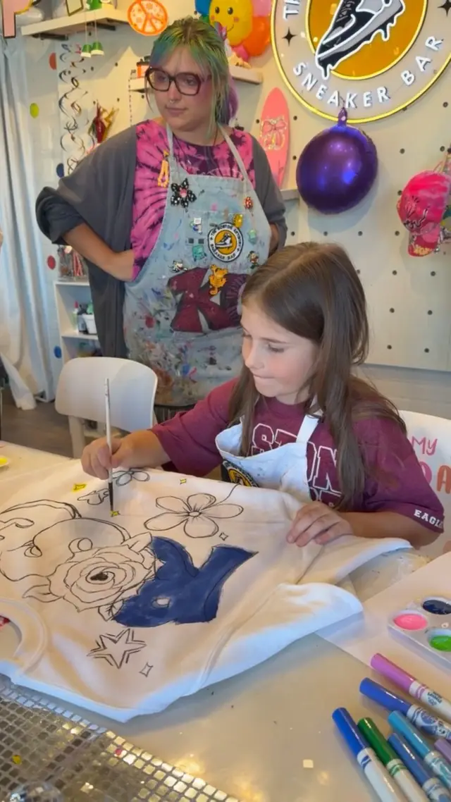 A girl paints a large blue letter on a white sweatshirt while a staff member in a studio apron watches