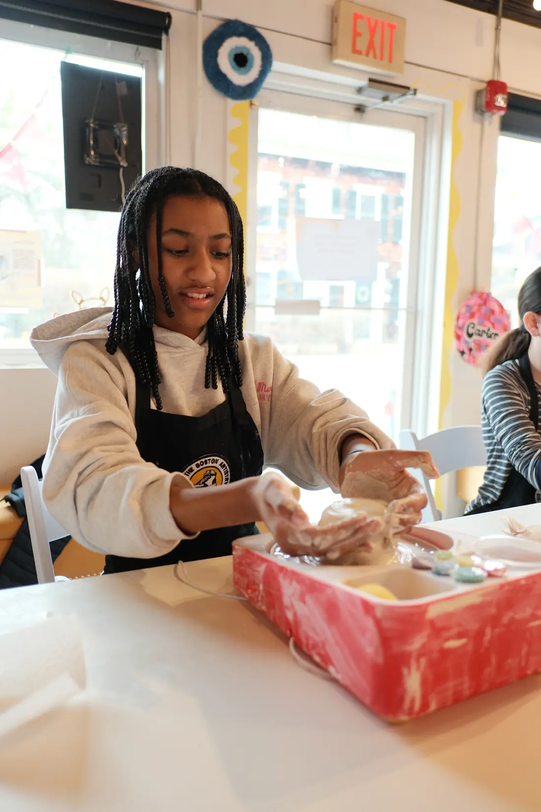 A girl in a studio apron shapes wet clay with both hands on a small tabletop pottery wheel at a white table