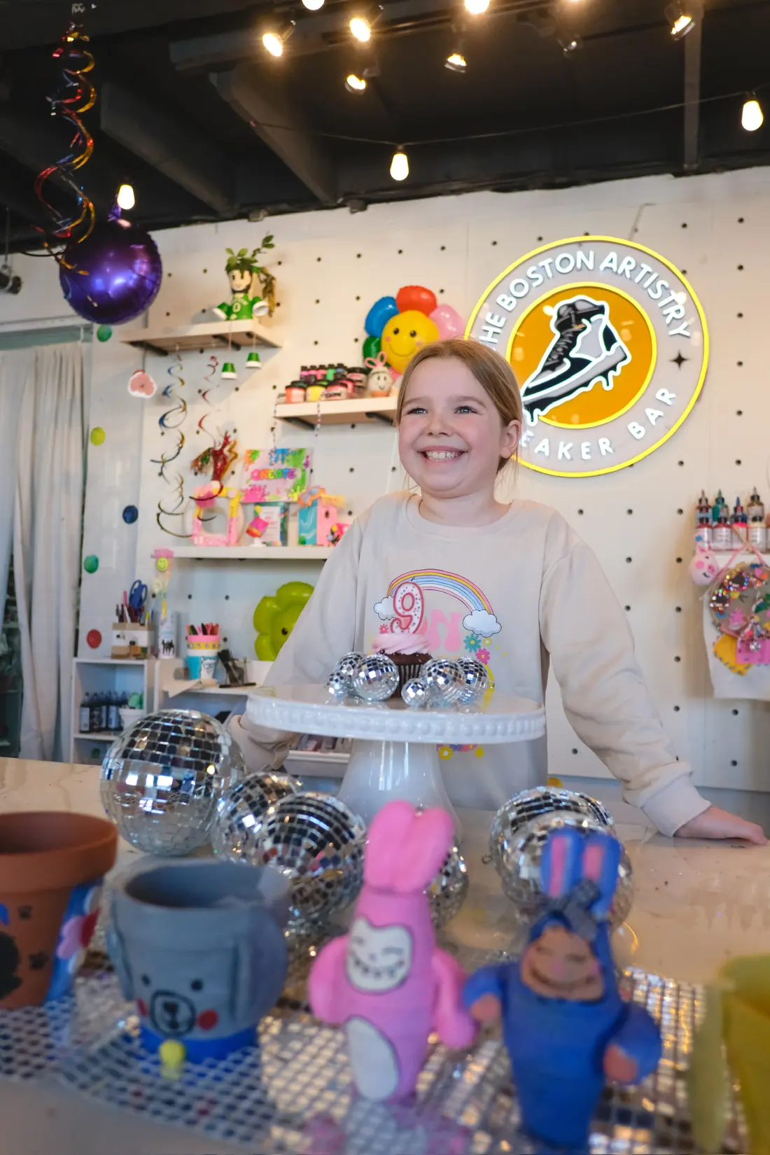 A girl in a tiara blows out candles on a stand of cupcakes under the neon studio sign