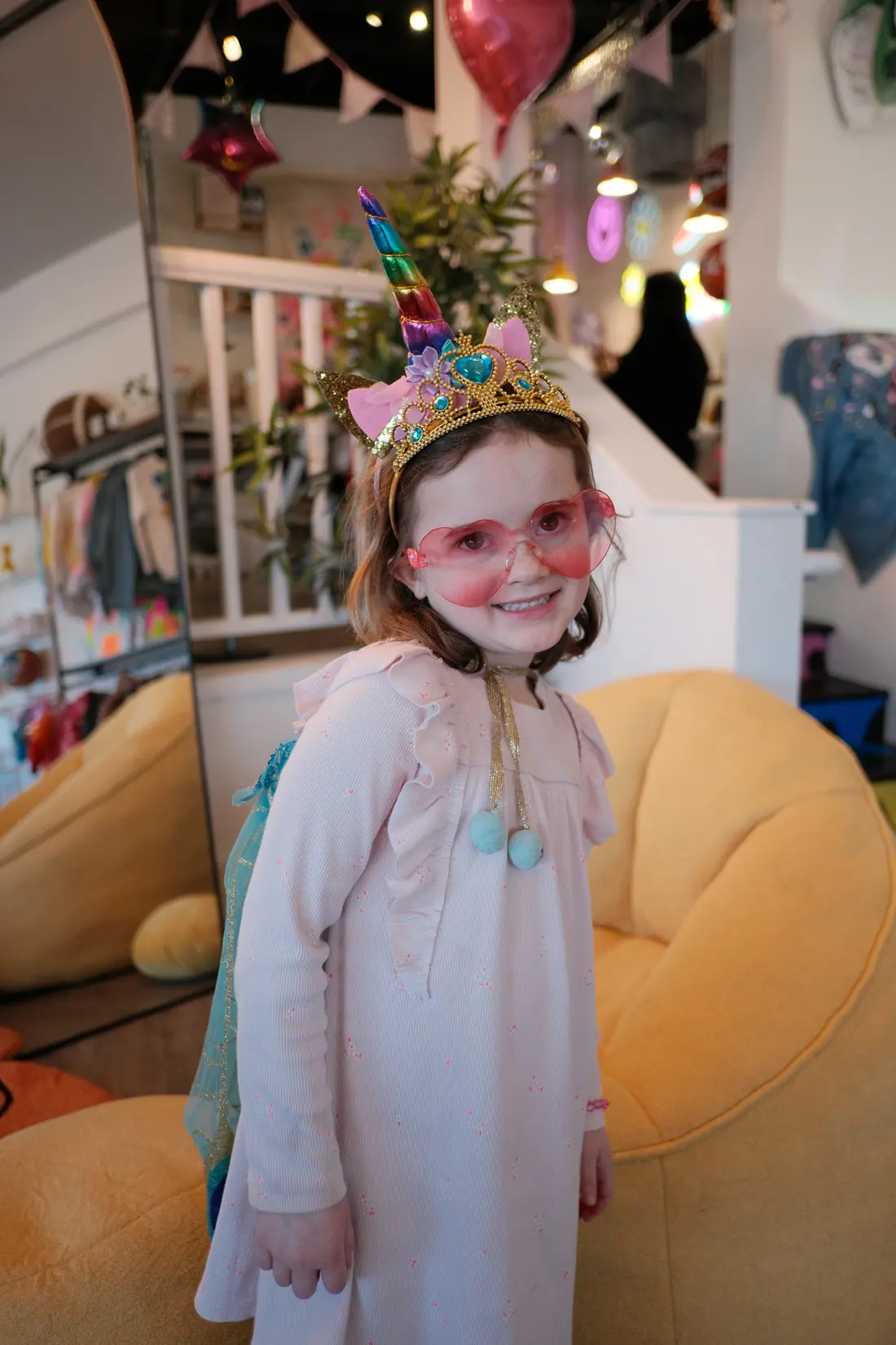 A small girl in pink heart glasses and a unicorn tiara smiles at her party