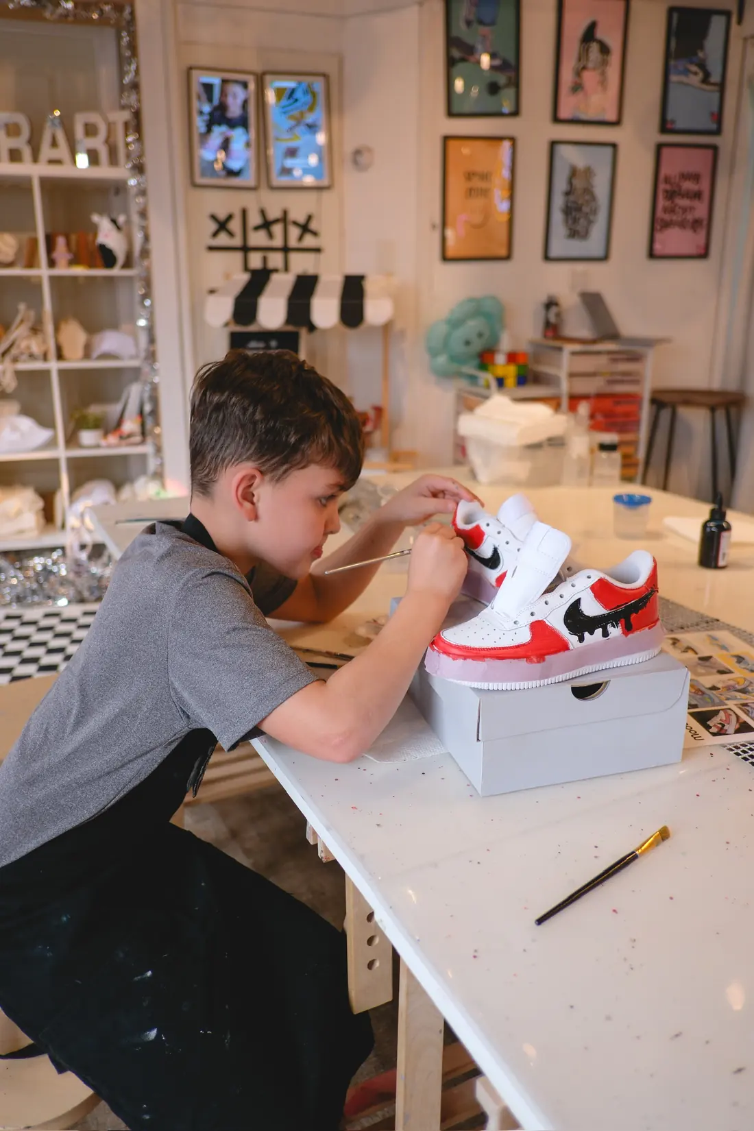 A boy in a black apron paints a white sneaker with red and black details beside its pair on a shoe box