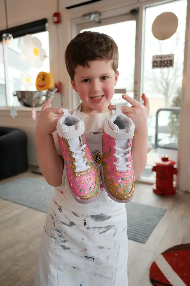 A smiling young boy in a paint splattered apron holds up sneakers painted pink and gold with sprinkles