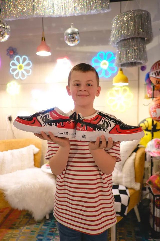 A boy in a red striped T-shirt holds out a pair of red, black and white custom painted sneakers