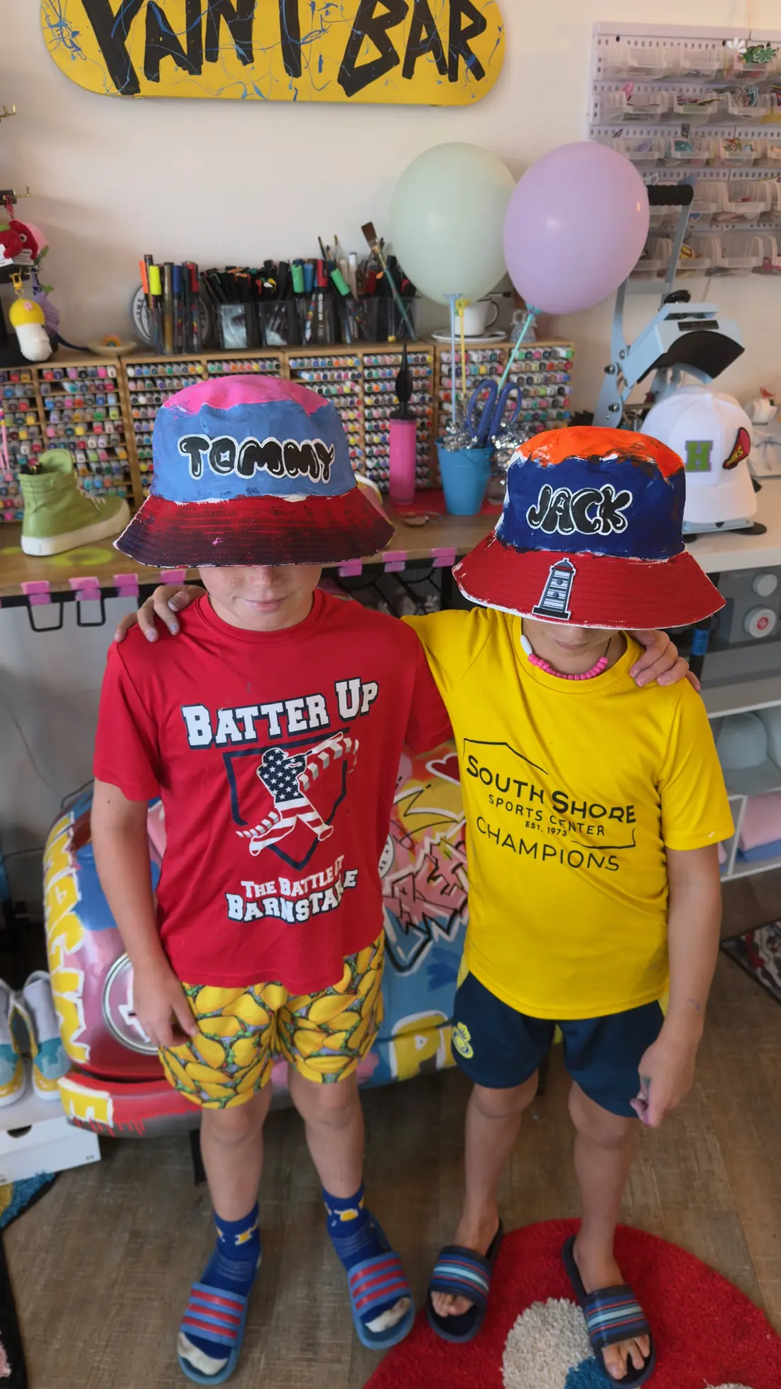Two boys stand arm in arm wearing hand painted bucket hats, brims pulled low over their eyes