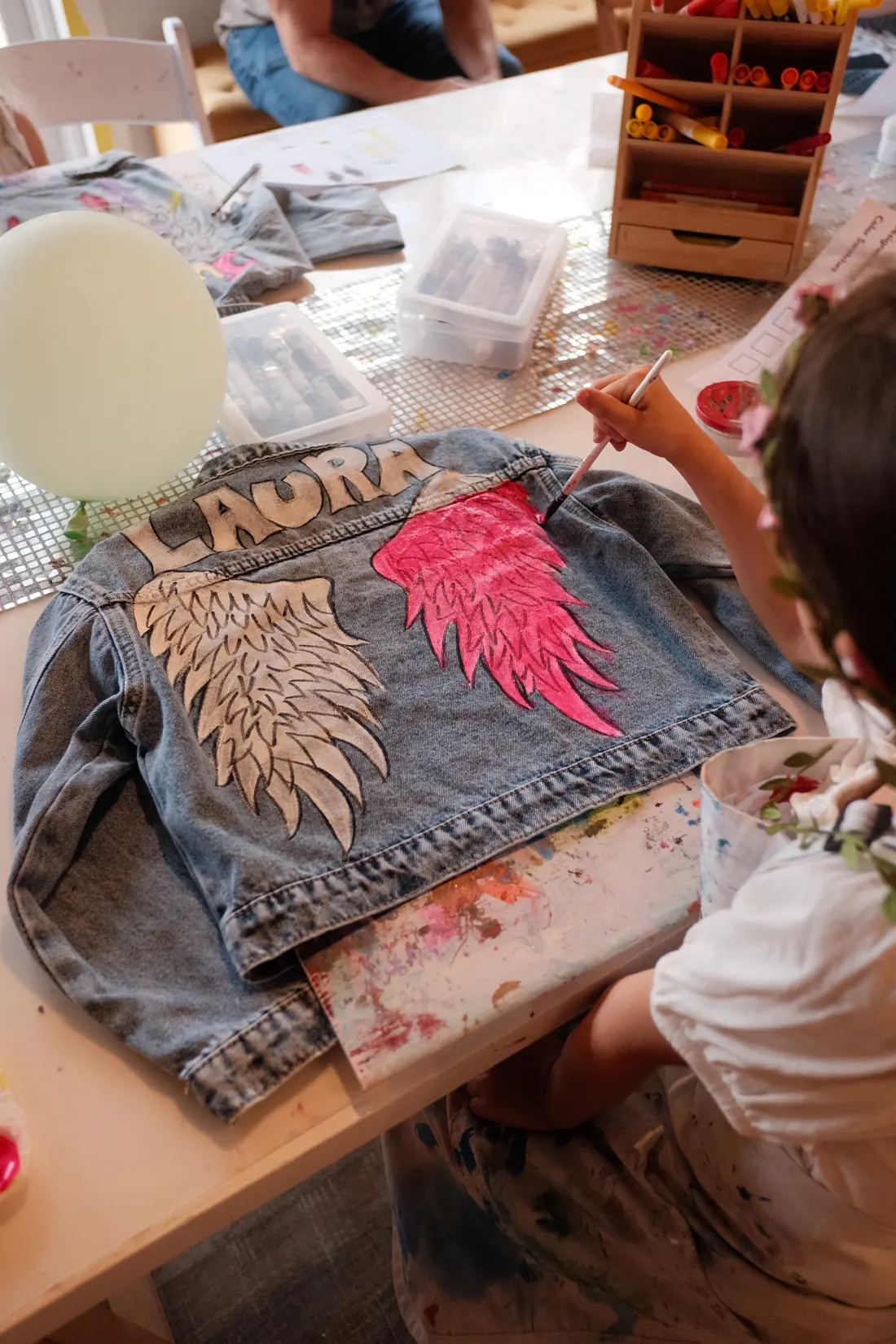 Seen from above, a child in a flower crown paints a pink wing on a denim jacket lettered with a name