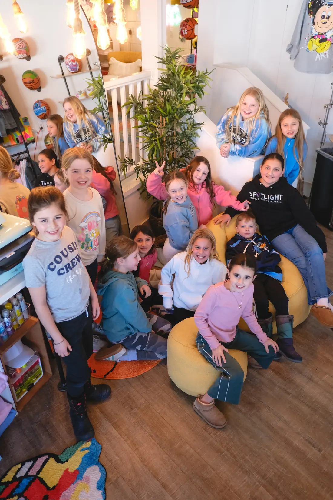 A large group of smiling children gathers on yellow bean bags beside the studio stairs, photographed from above