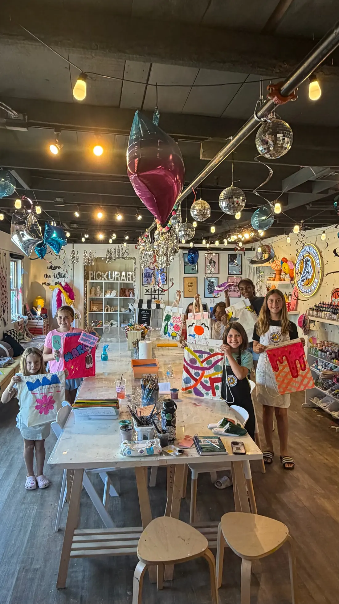 Children hold up tote bags they painted at a party table in the studio