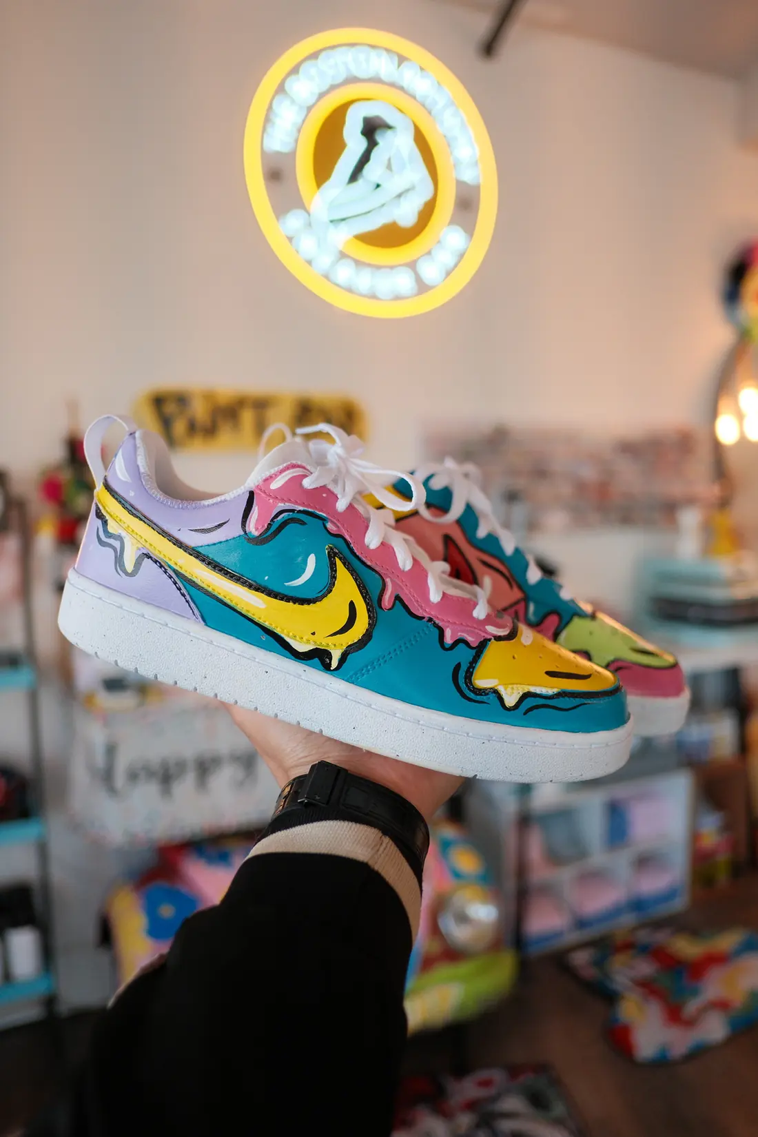An adult hand holds up a pair of low top sneakers painted in teal, pink and yellow drips inside the studio