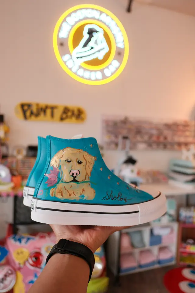 A teal custom painted high top sneaker with a golden retriever portrait, held up under the studio neon sign