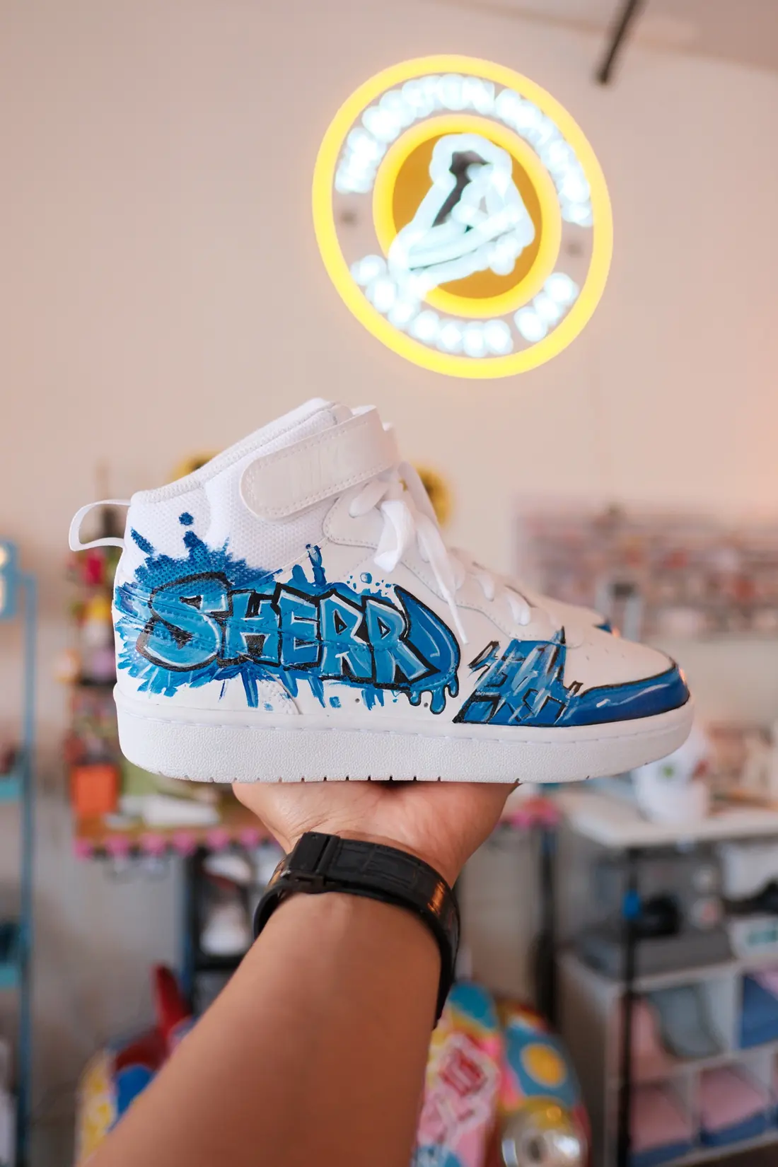 An adult hand holds a white mid top sneaker painted with blue graffiti lettering beneath the glowing neon studio sign