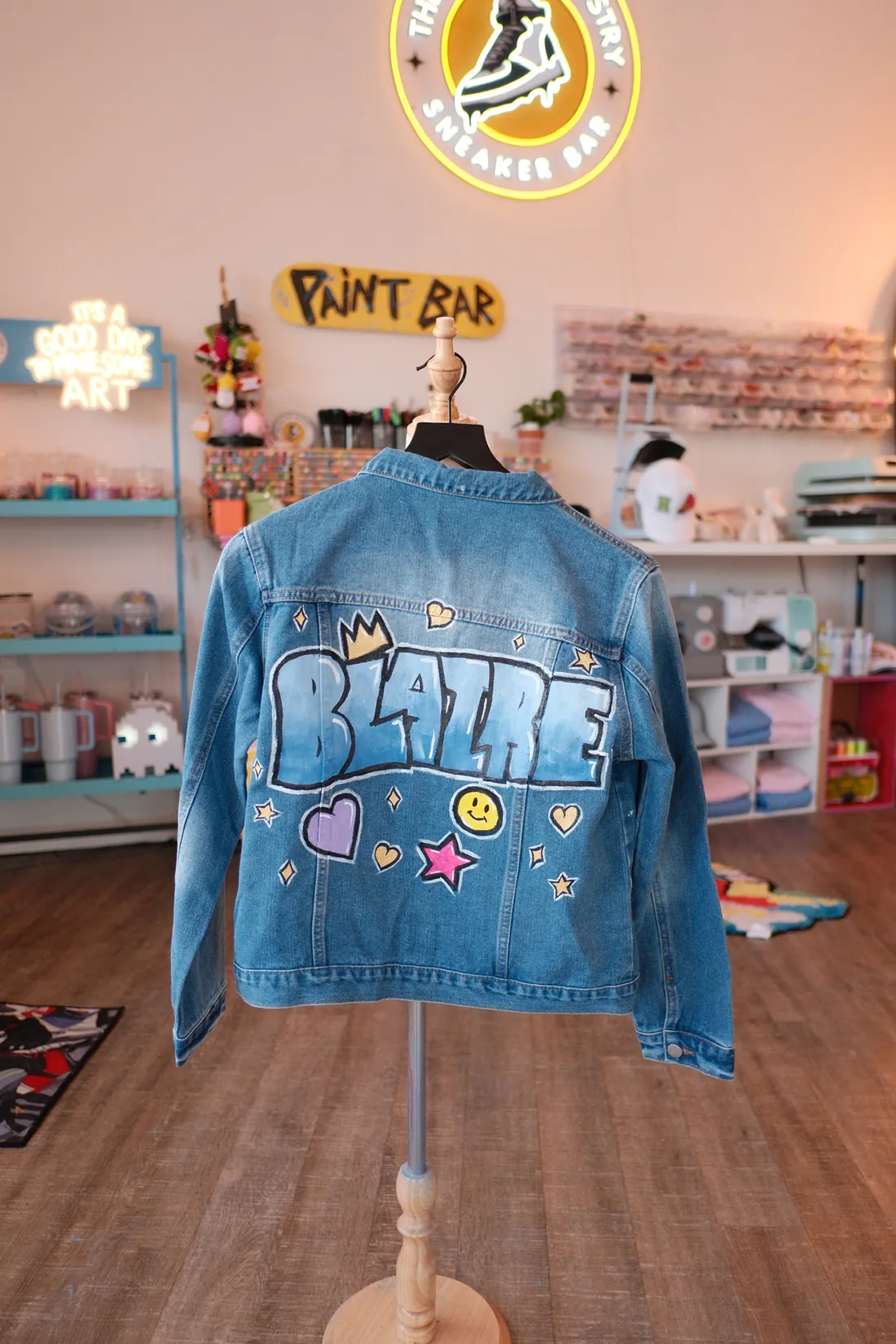 A denim jacket painted with a name in blue bubble letters, a crown, a smiley face and hearts hangs in the studio