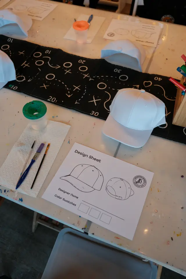 A blank white cap, a printed design sheet, three brushes and a water cup laid out on a paint speckled table