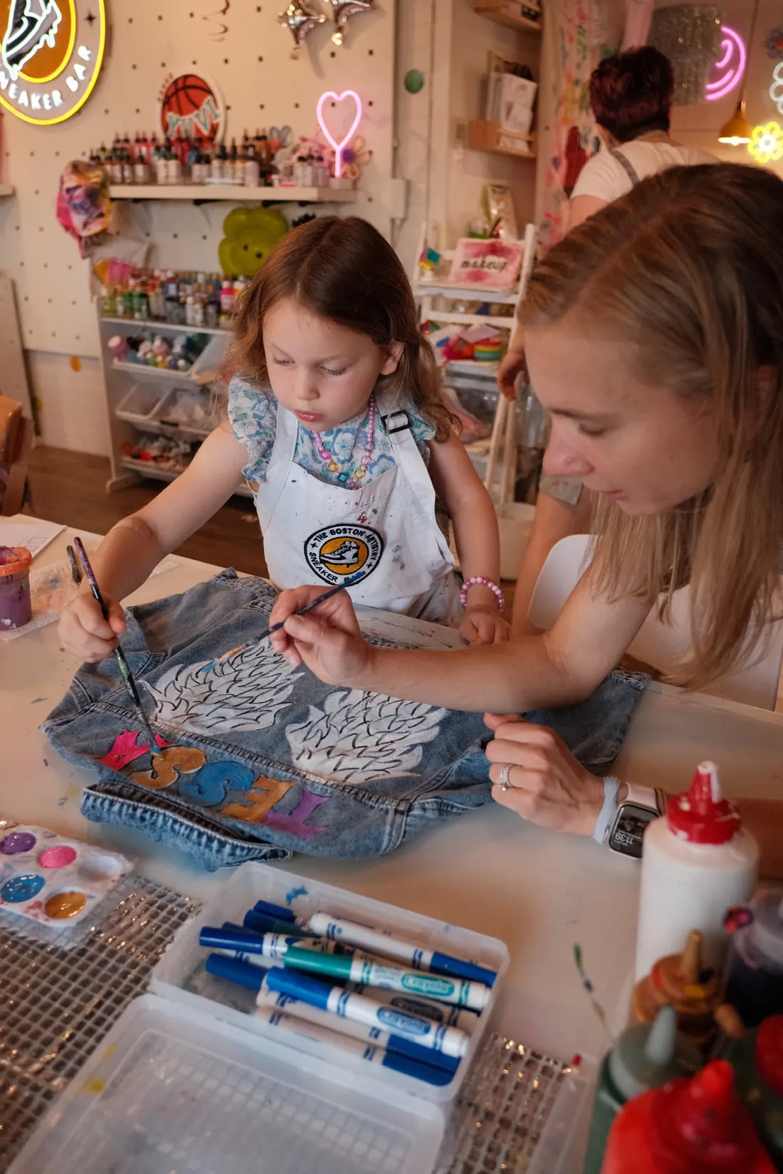 A small girl in a studio apron and a woman paint white wings and letters on a denim jacket together at the Hingham studio