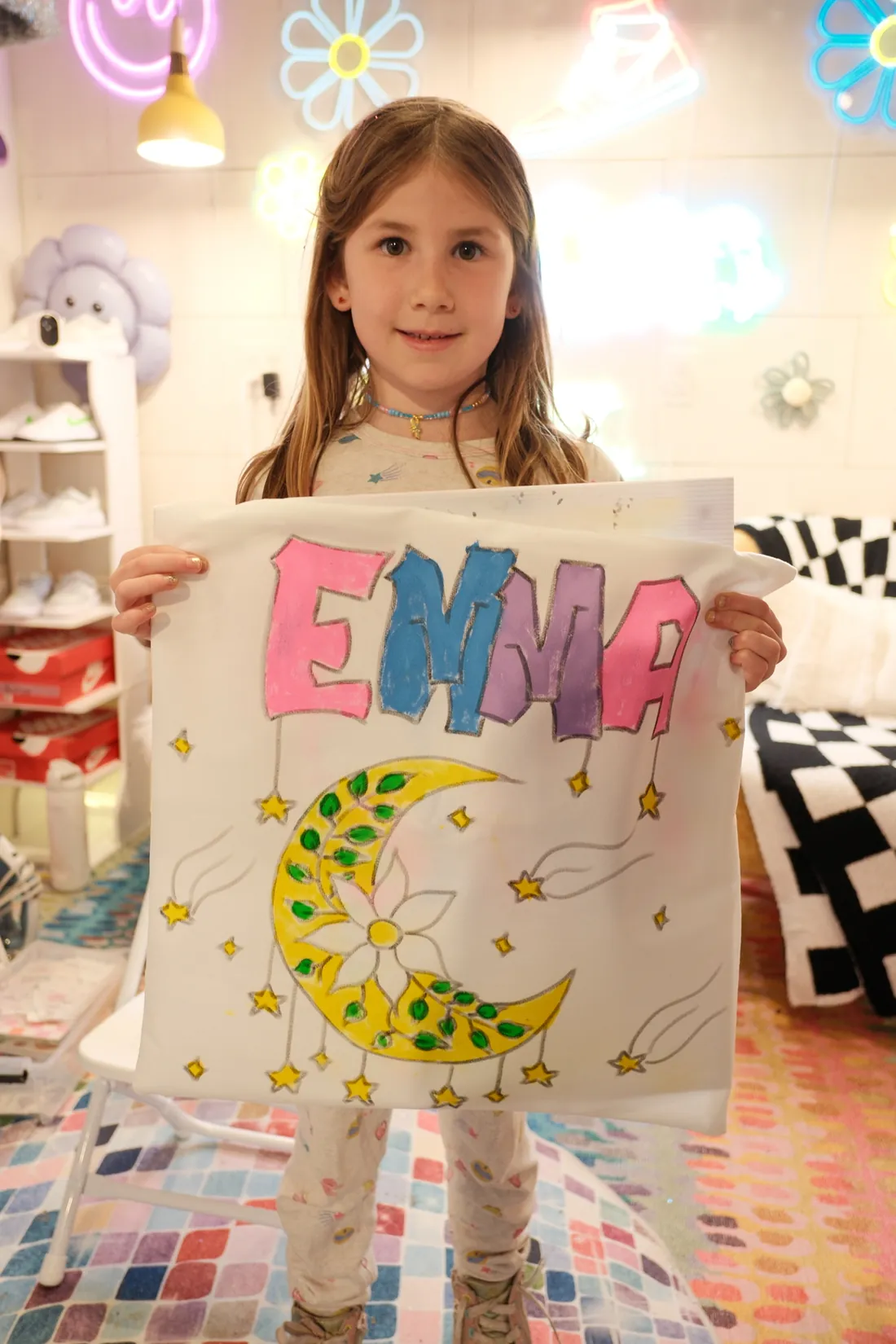 A girl holds up a white fabric square she painted with her name, a yellow moon and stars
