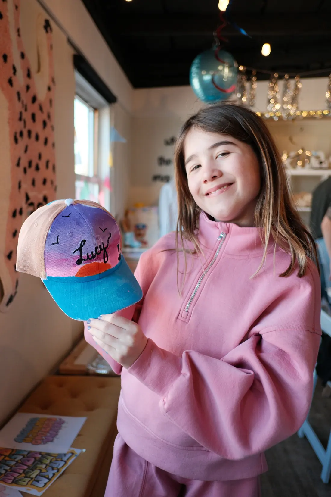 A smiling girl in a pink sweatshirt holds up a cap hand painted with a sunset sky and a name in script