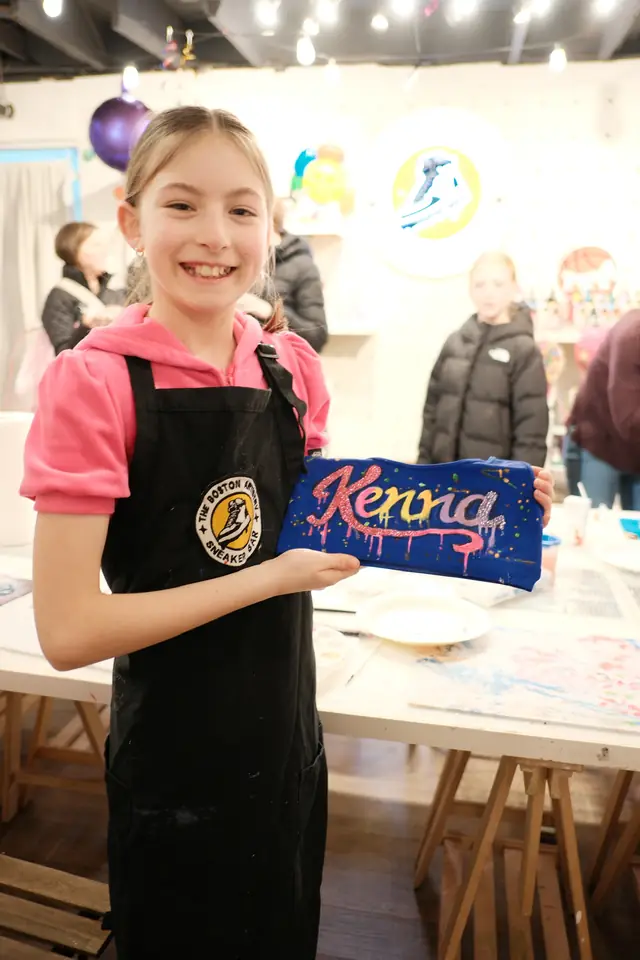 A smiling girl in a pink hoodie and studio apron holds a blue zip pouch painted with her name in glitter
