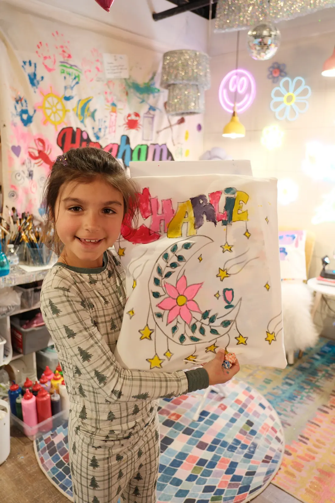 A smiling girl in plaid pajamas holds up a white fabric square she painted with her name, a leafy moon and stars