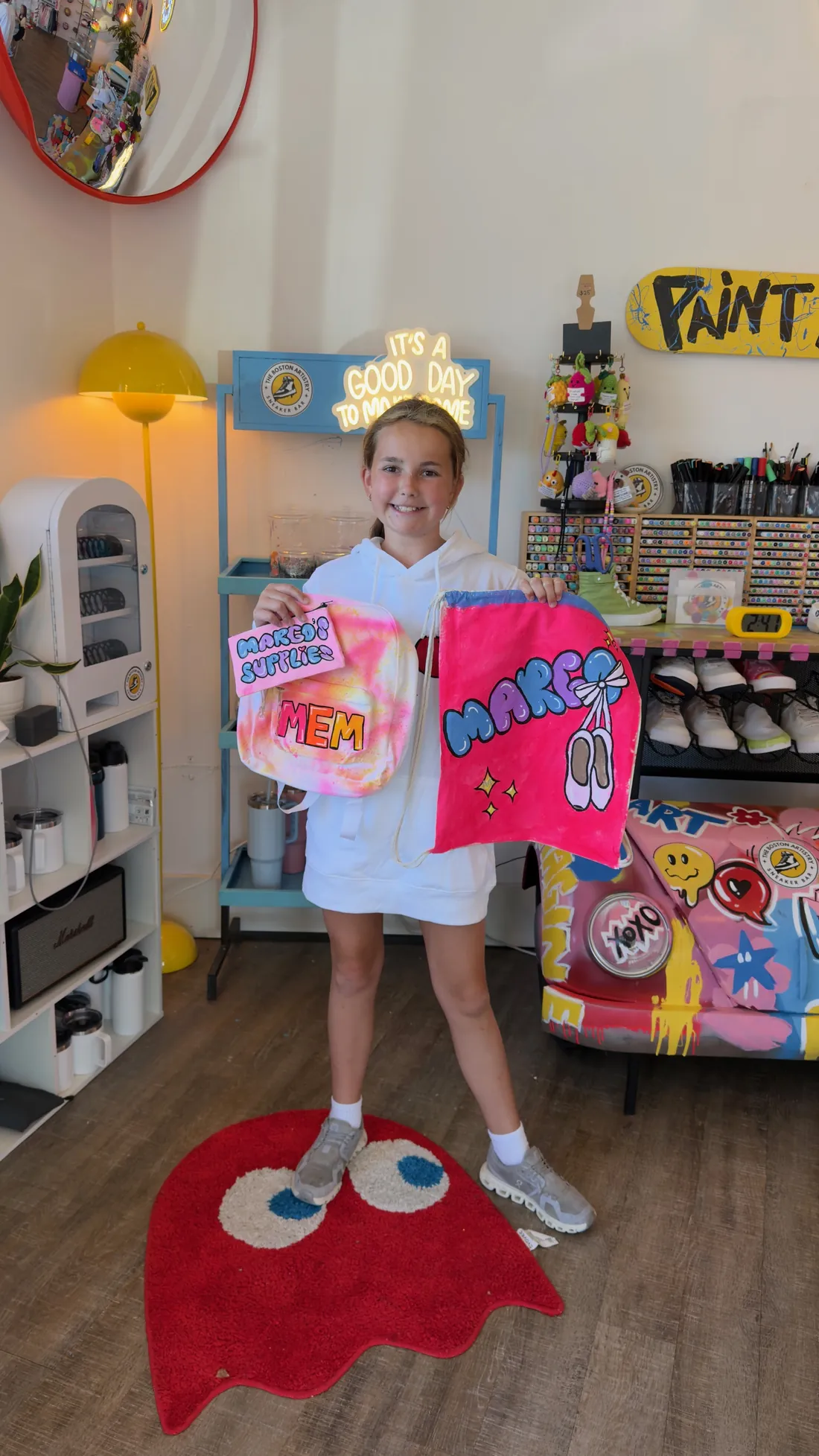A smiling girl shows a tie dye pouch and a pink drawstring bag painted with ballet shoes