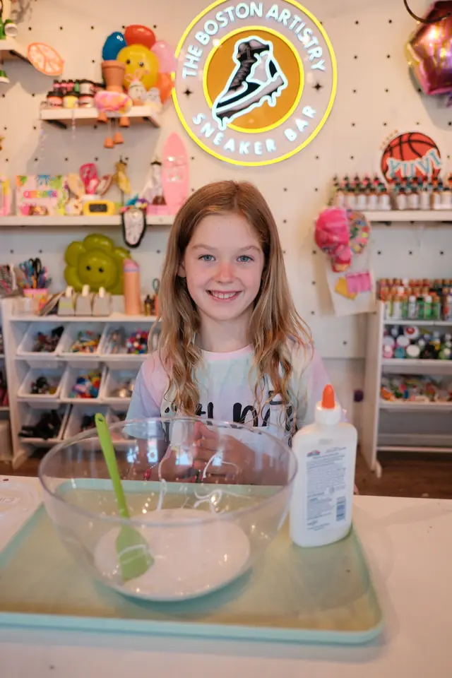 A smiling girl stands behind a mixing bowl beneath the neon sign of The Boston Artistry Sneaker Bar