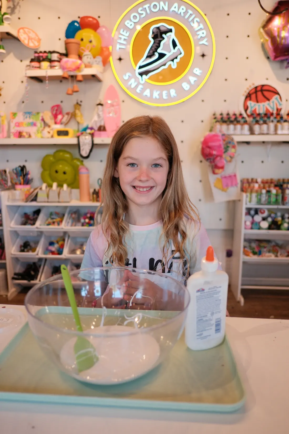 A smiling girl with long wavy hair stands behind a bowl of glue and a spatula beneath the neon studio sign