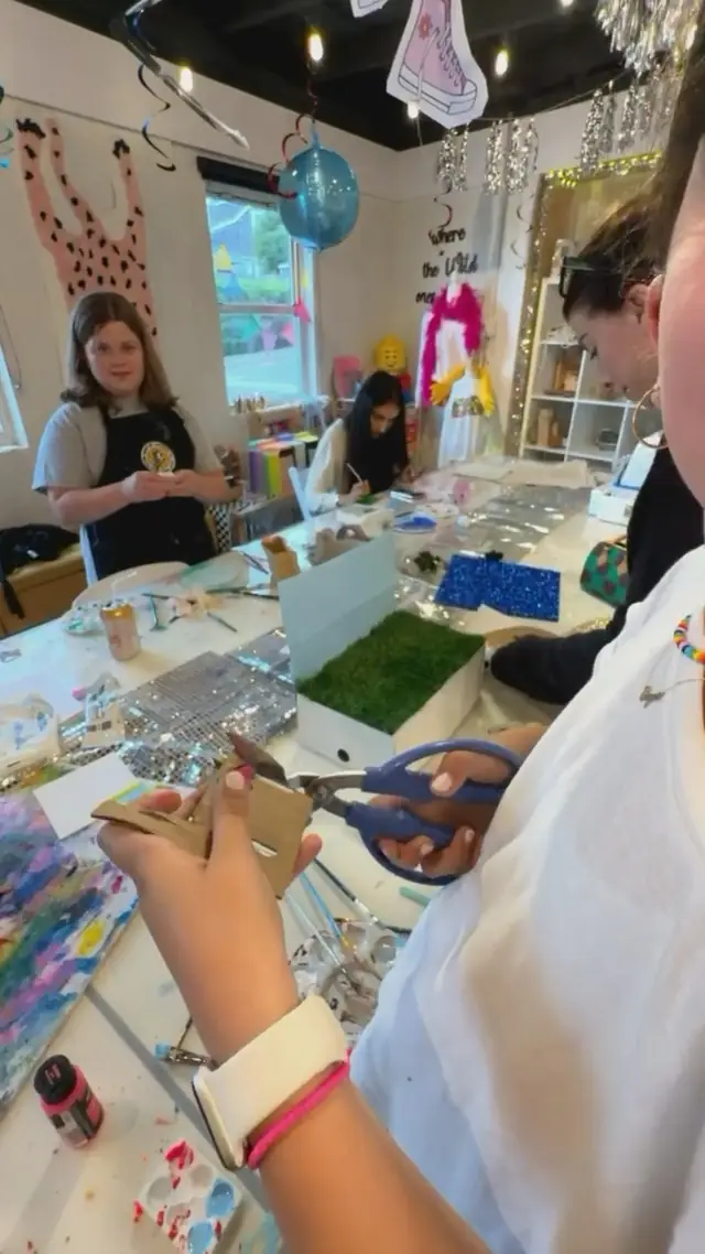 Tweens, teens and adults make mixed media projects around the long studio table while a woman cuts card with scissors