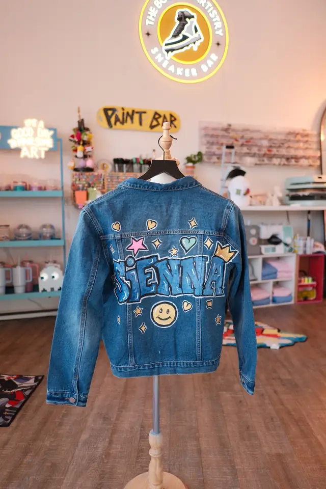 A dark denim jacket on display, hand painted with a name in blue letters, a gold crown, stars and a smiley face
