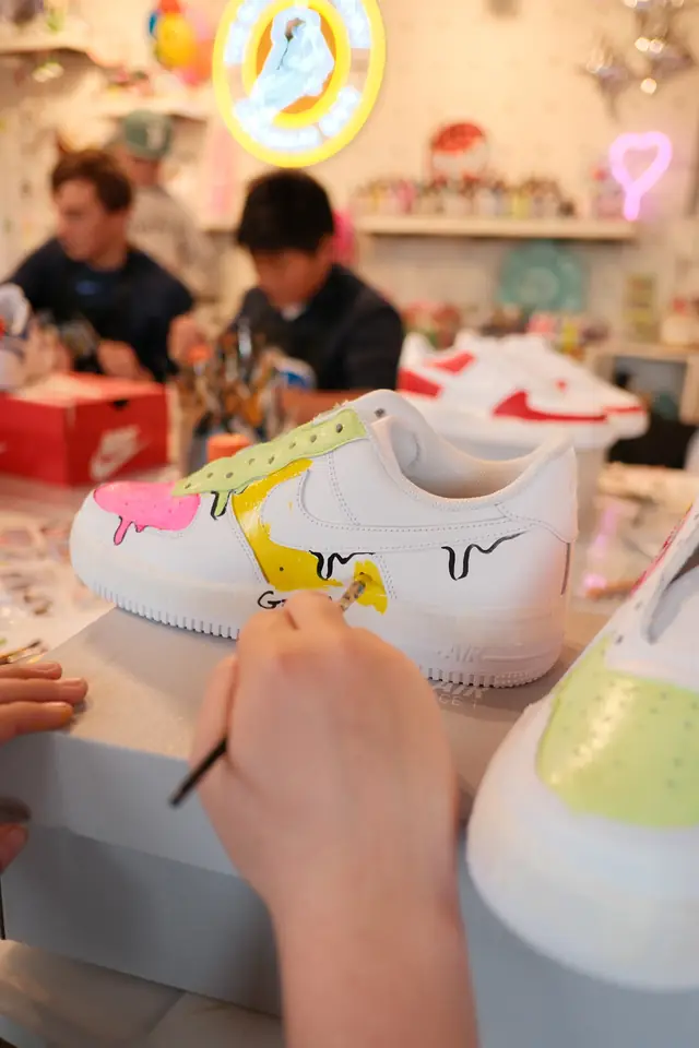 A hand paints black drip outlines onto a white sneaker with pink, green and yellow paint during a workshop