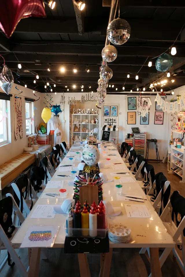 A long birthday party table set for cap painting with aprons, design sheets, paints and a Happy Birthday balloon centerpiece