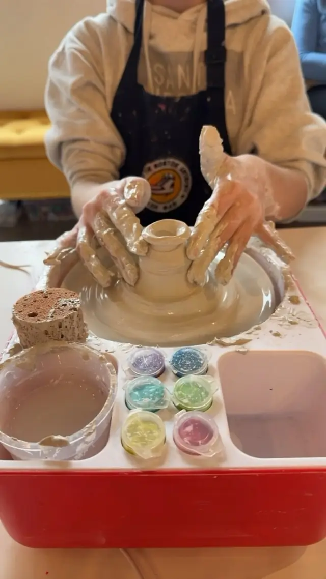 Clay covered hands in a studio apron shape a small pot on a tabletop pottery wheel