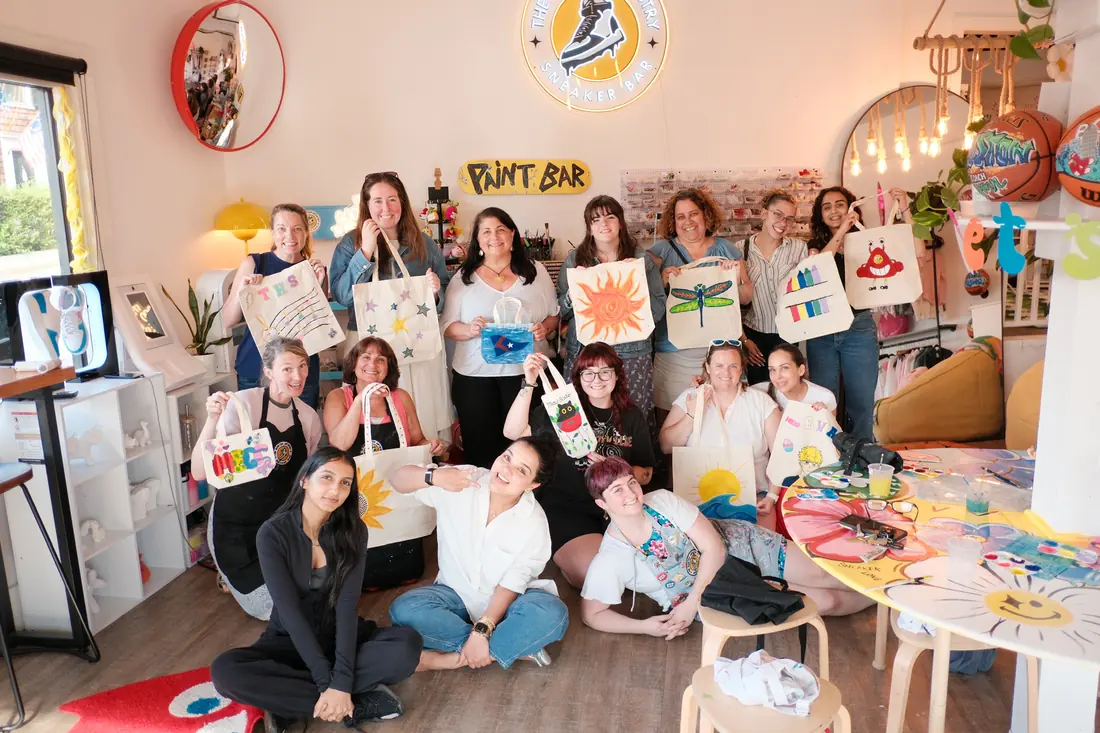 A group of about fifteen adults pose together in the studio holding up painted canvas tote bags beneath the neon sign