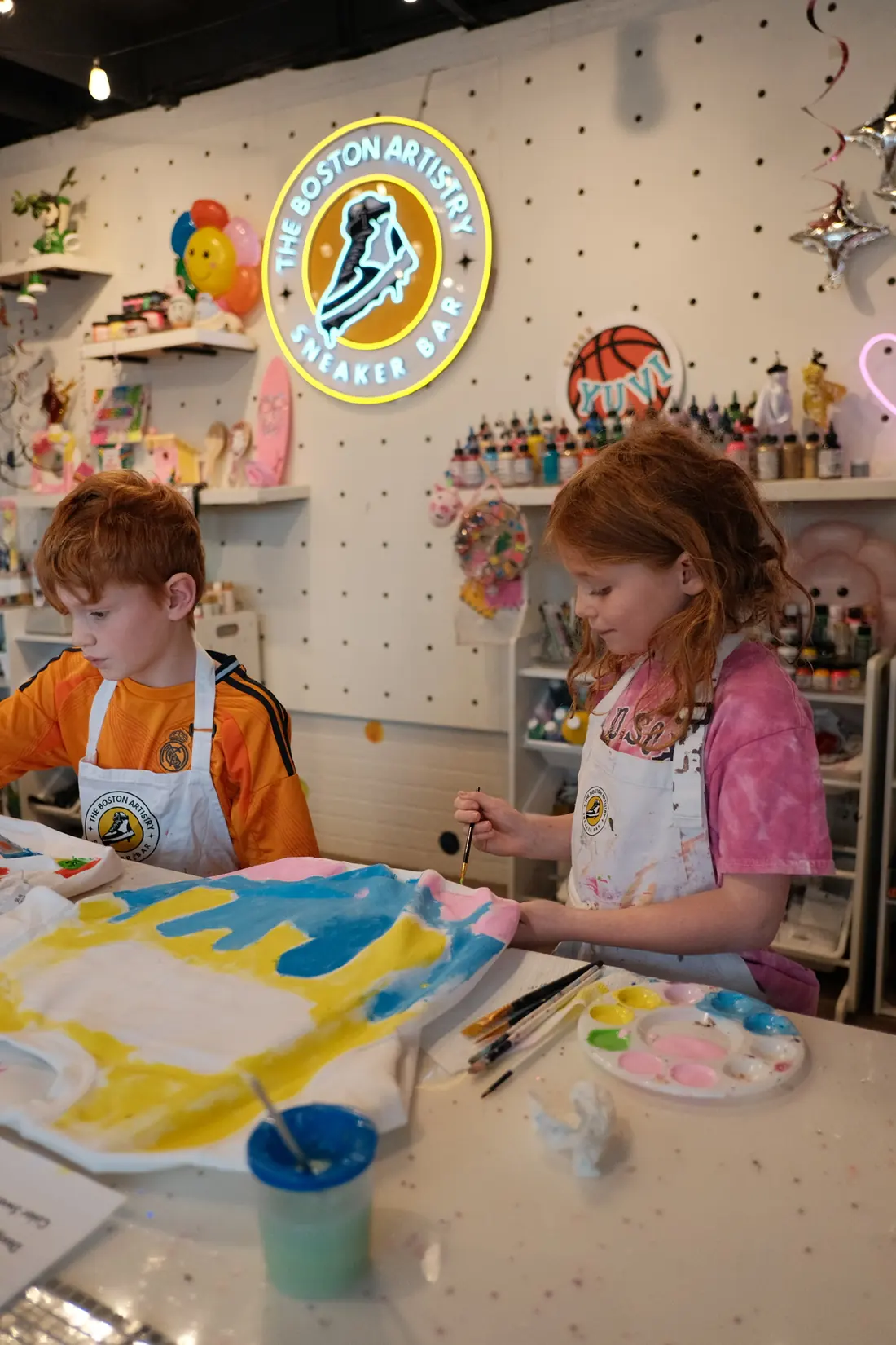 A boy and a girl in white aprons paint a white T-shirt yellow, blue and pink at the studio table