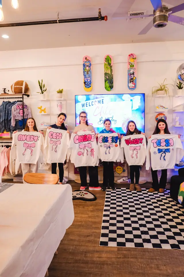 Six girls stand in a row in the studio, each holding up a white sweatshirt painted with her name and a bow