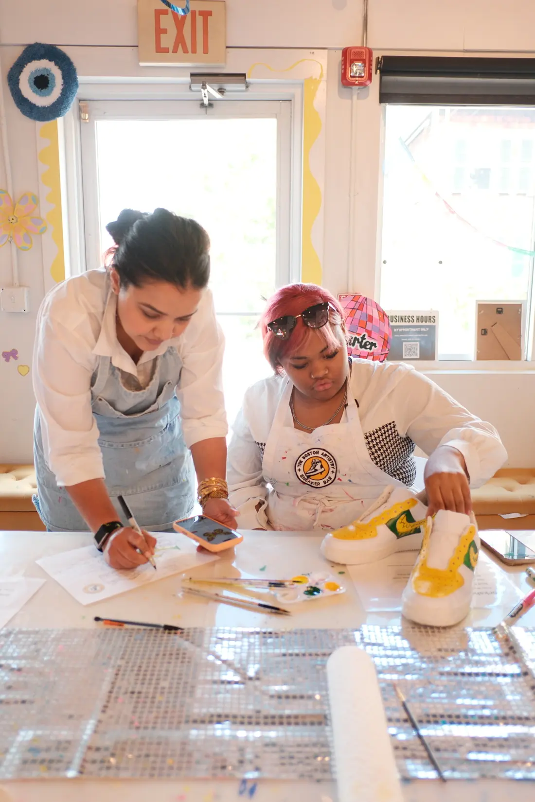A standing woman sketches on a design sheet while a seated adult paints white sneakers yellow and green