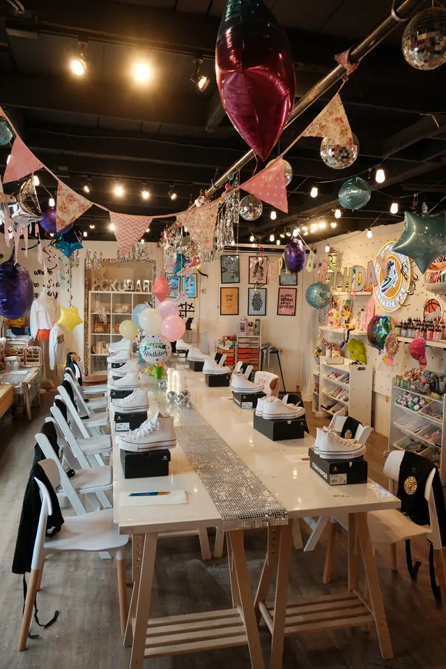 A birthday party table set with blank white high top sneakers on their boxes under balloons, bunting and disco balls