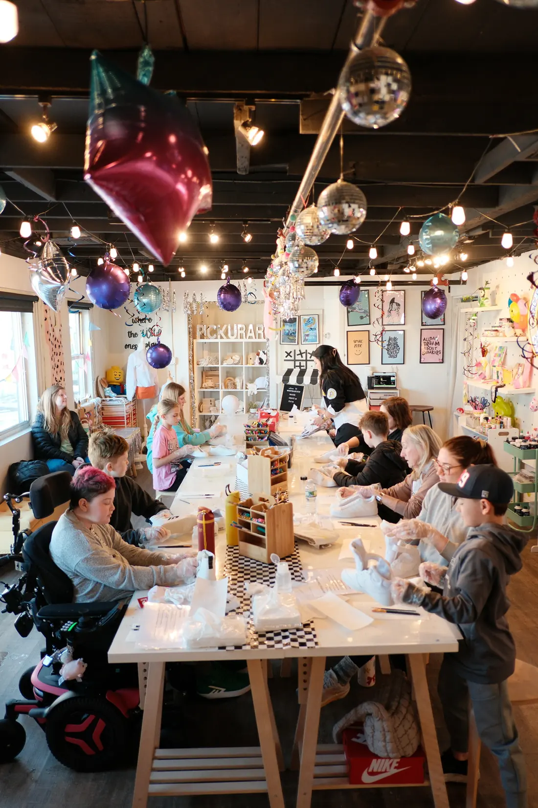 Children and adults sit along the long studio table preparing white sneakers under balloons, disco balls and string lights